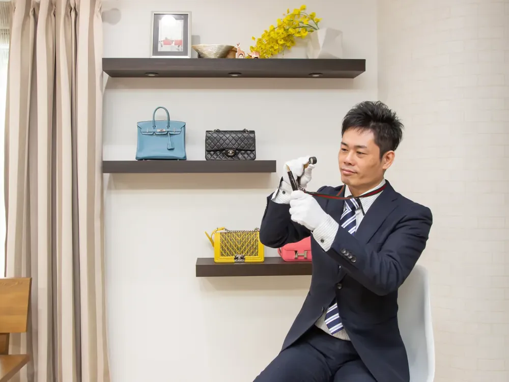 スーツ姿の鑑定士が、棚に並んだブランドバッグを見ながらルーペで確かめている写真