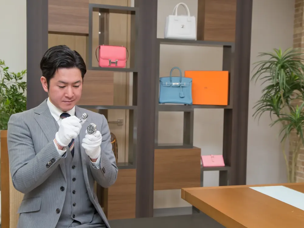 棚にエルメスのバッグが並ぶ店で、鑑定士がルーペで小物を確かめている写真