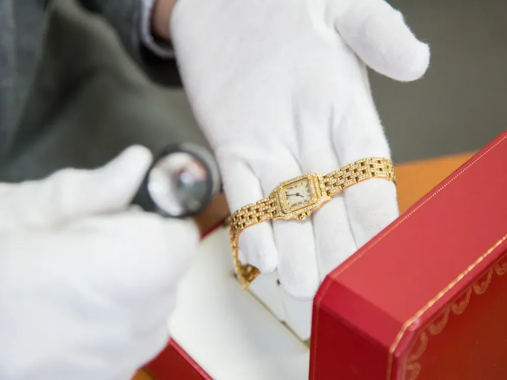 金色の腕時計を、白い手袋でルーペを持って確かめている写真。赤い箱が手前にある