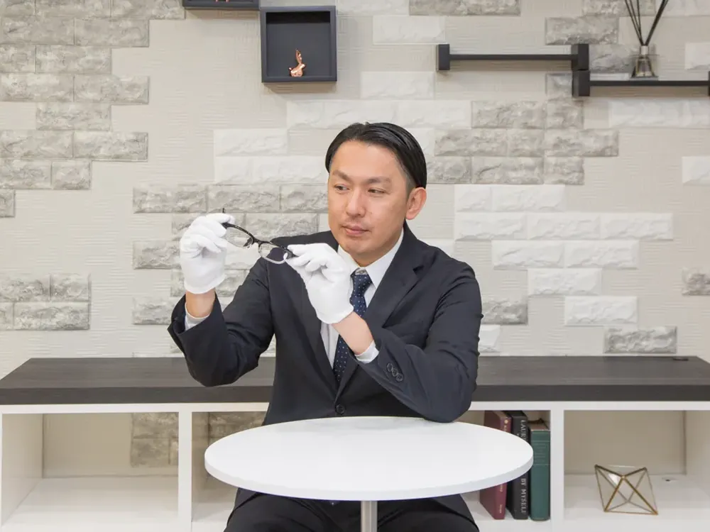 スーツ姿の鑑定士が、白い手袋でメガネのような品物を確かめている写真